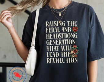 May include: Dark navy blue t-shirt with the text "RAISING THE FERAL AND HEADSTRONG GENERATION THAT WILL LEAD THE REVOLUTION" in white. The design includes red roses and a skull graphic. The Lucky Guppy Co. logo is in the bottom left.