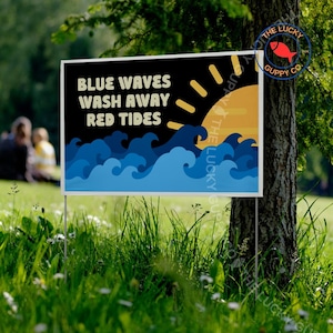 May include: A rectangular sign with the text "BLUE WAVES WASH AWAY RED TIDES" against a black background. The design includes blue waves and a yellow sun. The sign is placed in a grassy area.