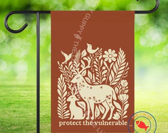 Könnte beinhalten: Eine braune Gartenflagge mit einer cremefarbenen Illustration eines Hirsches, eines Rehs, Vögeln und Blättern. Der Text "protect the vulnerable" steht unten. Die Flagge hängt an einem schwarzen Gartenstecker aus Metall.