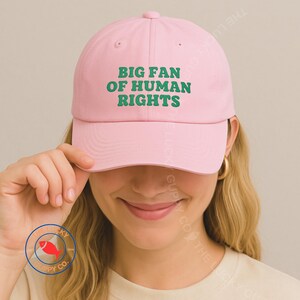 May include: A light pink baseball cap with the text "BIG FAN OF HUMAN RIGHTS" in green. The cap is made of a soft material and has a curved brim. The hat is a stylish accessory.