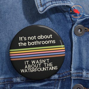 May include: A black button with white text that reads "It's not about the bathrooms. It wasn't about the waterfountains." The button has a rainbow stripe design.