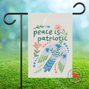 May include: A garden flag with the text "peace is patriotic" in green. The flag features a blue and green bird design with floral accents in pink and blue. The flag is hanging on a black metal stand.