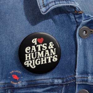 May include: A black badge with the words "I ❤️ CATS & HUMAN RIGHTS" in white, and a red heart symbol. The badge is pinned to a blue denim jacket. The Lucky Guppy Co. logo is visible.