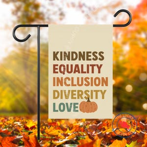 Può includere: Una bandiera da giardino beige con le parole "KINDNESS, EQUALITY, INCLUSION, DIVERSITY, LOVE" in lettere marroni e arancioni. Una grafica di zucca è sotto la parola "DIVERSITY". La bandiera è appesa a un supporto di metallo nero.