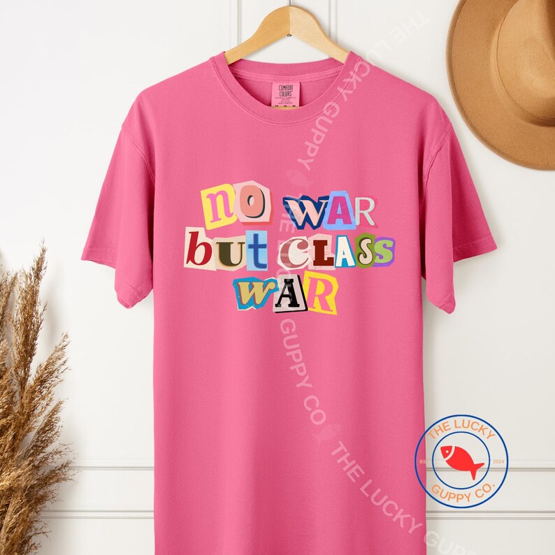May include: A bright pink t-shirt with the text "NO WAR but CLASS WAR" in a collage style. The text is in various colors and fonts. The shirt is hanging on a wooden hanger.