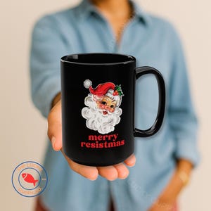 May include: Black ceramic mug featuring a vintage Santa Claus illustration with a red hat and white beard. The mug has the text "merry resistmas" in red. A festive holiday-themed mug.