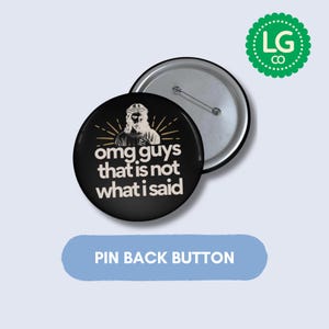 May include: A circular pin back button featuring a black background with a white illustration of a figure with a halo and the text "omg guys that is not what i said" in white lettering. The back of the button is silver with a metal pin mechanism.