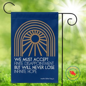 May include: A navy blue garden flag with a gold geometric design and the quote "We must accept finite disappointment but will never lose infinite hope" by Martin Luther King Jr. The flag is displayed on a black metal stand.