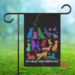 May include: Black garden flag featuring colorful dog silhouettes and paw prints. The text "it's okay to be different" is displayed at the bottom. The flag is hanging on a black metal garden stake.