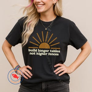 May include: Black t-shirt featuring a brown and gold sunburst graphic and the white text "build longer tables not higher fences". The shirt is a casual style.