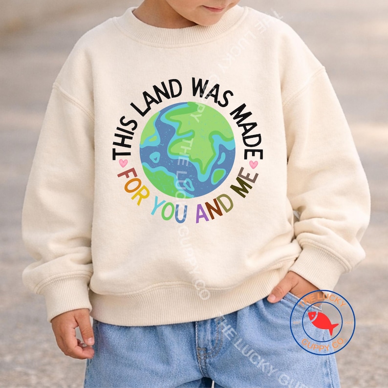 K&ouml;nnte beinhalten: Cremefarbenes Sweatshirt mit dem Text "This land was made for you and me", der einen Globus in Gr&uuml;n und Blau umgibt. Das Sweatshirt wird mit einer blauen Jeans kombiniert.