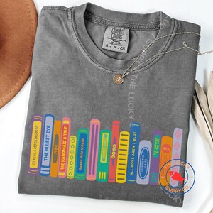 May include: A charcoal gray t-shirt featuring a graphic of colorful book spines with titles including To Kill a Mockingbird, The Bluest Eye, 1984, The Handmaid's Tale, Brave New World, Gender Queer, All Boys Aren't Blue, Looking for Alaska, and Tricks.