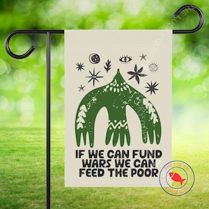 May include: A beige garden flag with a green graphic and the text "IF WE CAN FUND WARS WE CAN FEED THE POOR." The graphic includes stars, eyes, and a stylized bird. The flag is hanging on a black metal stand.
