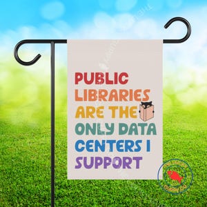 May include: A rectangular garden flag hanging on a black metal stand. The flag is off-white with multi-colored text that reads: PUBLIC LIBRARIES ARE THE ONLY DATA CENTERS I SUPPORT. A small black cat illustration is holding an open book.