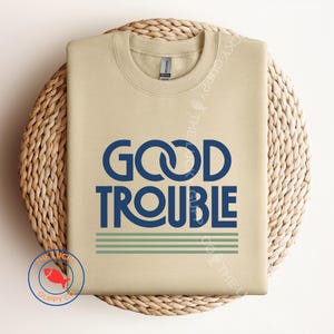 May include: A beige sweatshirt with the words "GOOD TROUBLE" in navy blue, with two green stripes below the text. The sweatshirt is displayed on a woven, circular surface.