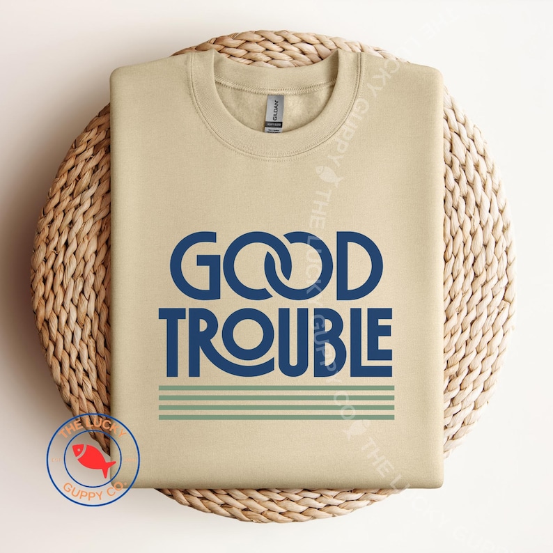 May include: A beige sweatshirt with the words "GOOD TROUBLE" in navy blue, with two green stripes below the text. The sweatshirt is displayed on a woven, circular surface.