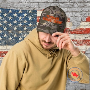 May include: A camouflage baseball cap with the text "Veterans Against Trump" in orange lettering. The cap is being worn by a person with a beard and a tan hoodie.
