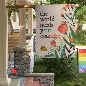May include: A beige garden flag with the text "the world needs your courage" in black. The flag features floral illustrations in orange, yellow, and green. A smaller rainbow flag with peace symbols is also visible.