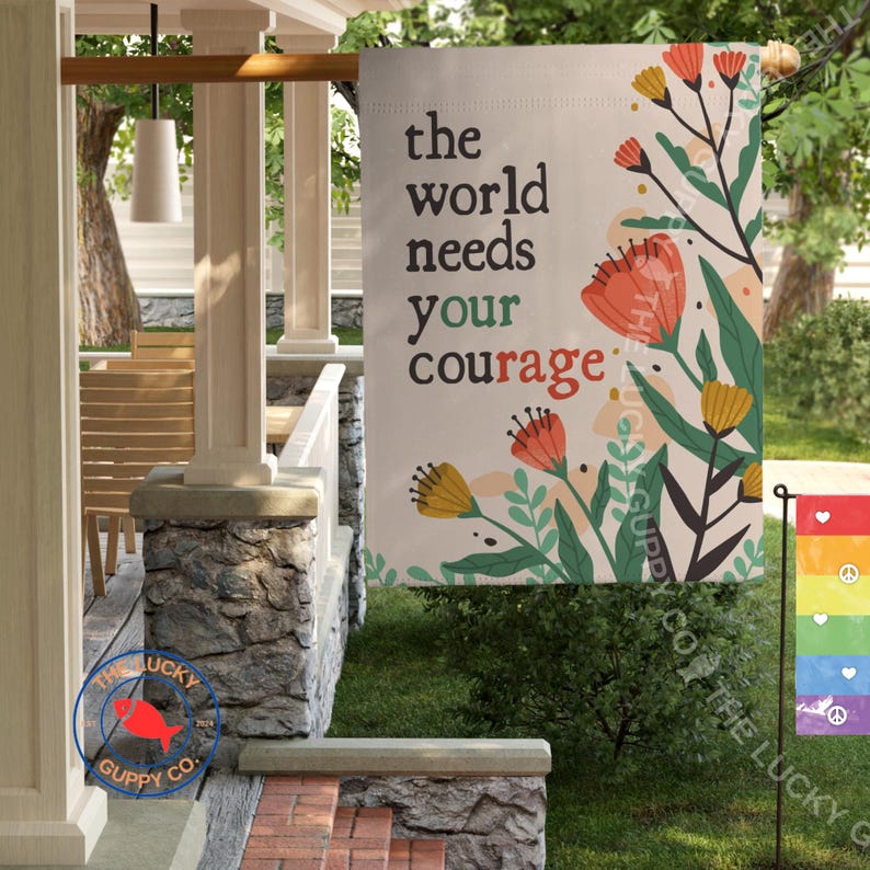 May include: A beige garden flag with the text "the world needs your courage" in black. The flag features floral illustrations in orange, yellow, and green. A smaller rainbow flag with peace symbols is also visible.