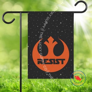 May include: A black garden flag with an orange emblem and the word "RESIST" in white. The flag has a starry background and is displayed on a black metal stand. The flag is designed for outdoor use.
