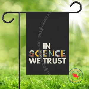 May include: A black garden flag with the text "IN SCIENCE WE TRUST" in colorful letters. The flag is hanging from a black metal stand, with a green grassy background.