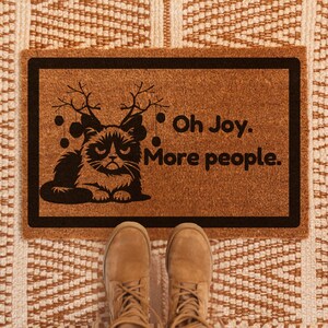 May include: A brown doormat with a black cat wearing reindeer antlers and the text "Oh Joy. More people."