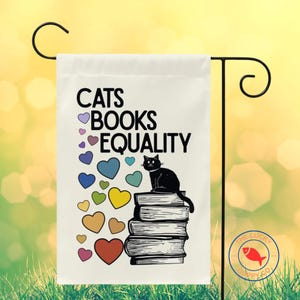 May include: A white garden flag with the words "CATS BOOKS EQUALITY" in black. A black cat sits atop a stack of books, with colorful hearts surrounding the text. The flag is displayed on a black metal stand.