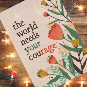 May include: A white tote bag with the text "the world needs your courage" in dark brown. The bag features floral illustrations in orange, yellow, and green. The Lucky Guppy Co. logo is visible in the bottom left corner.