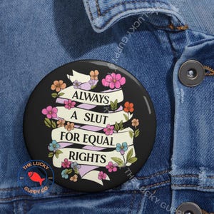 May include: A black circular badge with the slogan "ALWAYS A SLUT FOR EQUAL RIGHTS" in white, with floral decorations. The design features a banner and colourful flowers. The Lucky Guppy Co. logo is at the bottom of the badge.