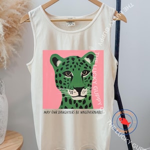 Könnte beinhalten: Ein cremefarbenes Tanktop mit einem Grafikdruck eines grünen Leoparden mit schwarzen Flecken vor einem rosa quadratischen Hintergrund. Unter dem Leoparden steht der Text "MAY OUR DAUGHTERS BE UNGOVERNABLE" in schwarzen Großbuchstaben. Rundhalsausschnitt.