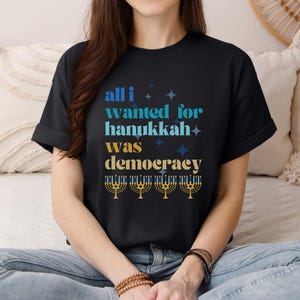May include: Black t-shirt with the text "all i wanted for hanukkah was democracy" in blue and yellow. The design includes star shapes and menorah illustrations. The shirt is a casual wear item.
