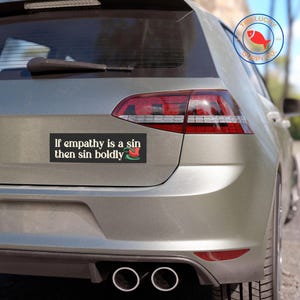 May include: A silver car with a black bumper sticker that reads "If empathy is a sin then sin boldly". The sticker has a red flower graphic on the right side.
