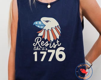 May include: Navy blue tank top featuring a patriotic eagle design with red, white, and blue accents. The text "Resist like it's 1776" is displayed below the eagle. A round logo with a red fish is visible on the bottom right.