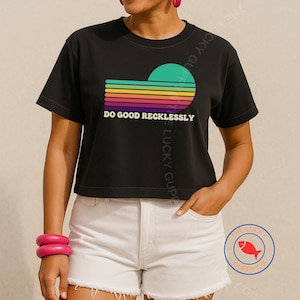 May include: Black crop top with a colorful retro sunset graphic and the text "DO GOOD RECKLESSLY". The graphic features a teal sun and horizontal stripes in yellow, orange, pink, purple, and blue. The shirt is paired with white shorts.