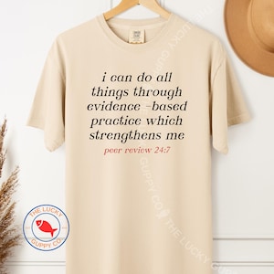 May include: A beige t-shirt with the text "i can do all things through evidence-based practice which strengthens me peer review 24:7" in black and red. The shirt has a crew neck and short sleeves. The Lucky Guppy Co. logo is visible on the bottom left.