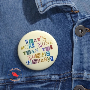 May include: A round, off-white button with the text "What's More Punk Than The Public Library?" in colorful, cut-out style letters. The button is pinned to a denim jacket. The Lucky Guppy Co. logo is visible.