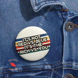 May include: A white button with the text "IT'S NOT RED VS. BLUE IT'S THE RICH VS. YOU" in a retro design. The button has blue, red, yellow, and green stripes. The button is on a denim jacket.