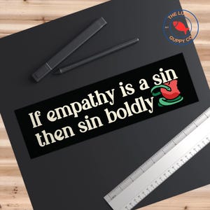 May include: Black and white sticker with the text "If empathy is a sin then sin boldly" in white font. A red and green snake with a red apple in its mouth is in the bottom right corner.