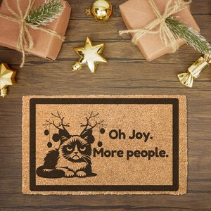 May include: A brown coir doormat with a black border and a black and white illustration of a grumpy cat wearing reindeer antlers. The text "Oh Joy. More people." is printed on the doormat.