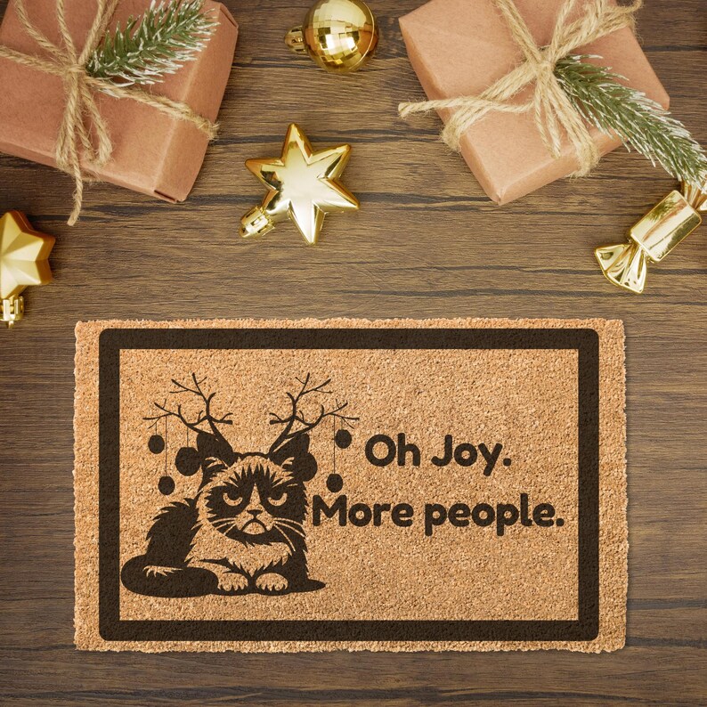 May include: A brown coir doormat with a black border and a black and white illustration of a grumpy cat wearing reindeer antlers. The text "Oh Joy. More people." is printed on the doormat.