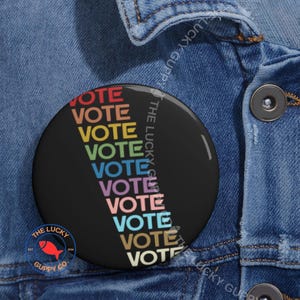 May include: A circular black badge with the word "VOTE" in rainbow colours repeated vertically. The badge also features the text "THE LUCKY GUPPY CO" and a red fish logo. The badge is set against a denim jacket.