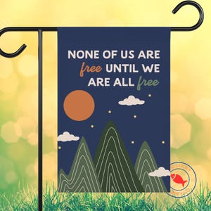 May include: A navy blue garden flag with the text "NONE OF US ARE free UNTIL WE ARE ALL free." The design includes an orange sun, white clouds, and green mountains. The flag is displayed on a black metal stand.