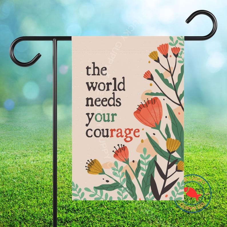 May include: A garden flag with the text "the world needs your courage" in black and red. The flag features floral illustrations in orange, yellow, and green against a beige background. The flag is hanging on a black metal stand.