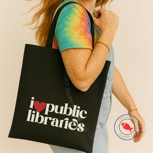 May include: Black tote bag with the text "i ❤️ public libraries" in white and a red heart. The bag is held by a person wearing a tie-dye shirt and denim overalls. A small logo with a red fish is visible.