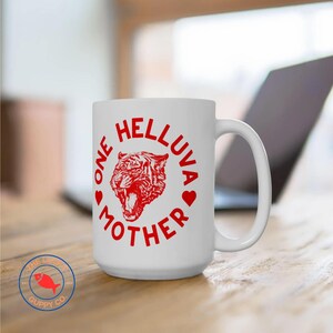 May include: White ceramic mug with a red graphic of a tiger's head inside a circle with the text "One Helluva Mother" around it.