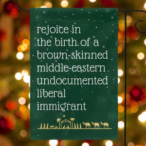 May include: A dark green garden flag with white text that reads "rejoice in the birth of a brown-skinned middle-eastern undocumented liberal immigrant." The bottom of the flag features a nativity scene.