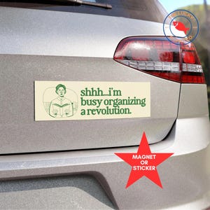 May include: A rectangular bumper sticker with the text "shhh...I'm busy organizing a revolution." The sticker features a green illustration of a person reading a book. A red star with the words "MAGNET OR STICKER" is below the bumper sticker.