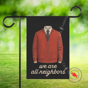 May include: A black garden flag features a graphic of a red sweater, white collared shirt, and striped tie. The text "we are all neighbors" is below the graphic. The flag is displayed on a black metal stand.
