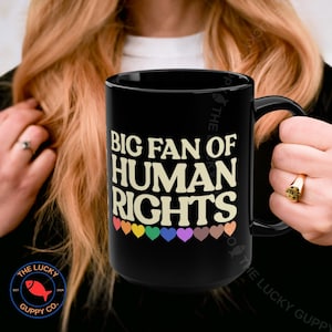 May include: Black ceramic mug with the text "BIG FAN OF HUMAN RIGHTS" in a bold, off-white font. Below the text are colorful hearts. The mug has a large handle and is a statement piece.