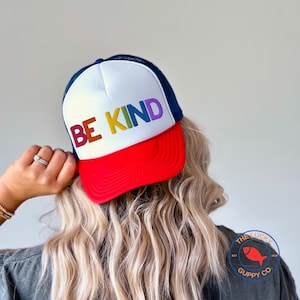 May include: A white and red trucker hat with a navy blue mesh back. The hat has the words "BE KIND" embroidered on the front in rainbow colors.